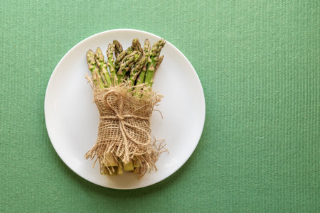 Asparagus shoots are tied with a jute rope on a white plate. Fresh asparagus on a green background.