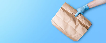 The concept of food delivery. A man's hand in medical gloves holds a paper bag against a blue...