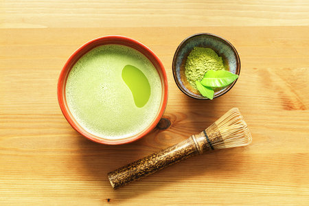 A bowl of matcha tea on a wooden table with a whisk and powder.
