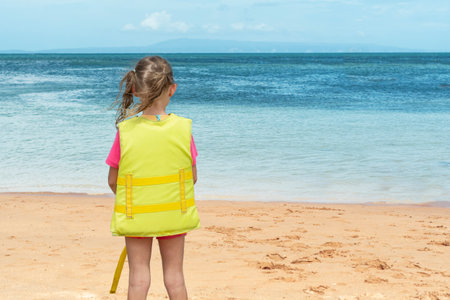 Little girl in yellow safety jacket standing in front of the ocean.の写真素材
