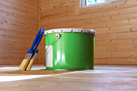 Green paint bucket with blue brushes on a wooden floor in a timber-clad room, representing home renovationの写真素材