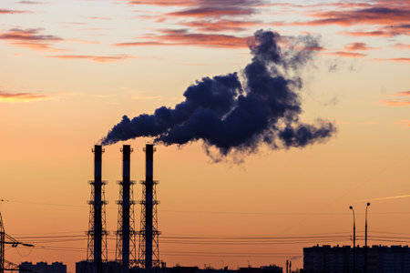 Factory chimneys release dense dark smoke into the air, colored by sunset hues. Industrial pollution of a metropolis.の写真素材