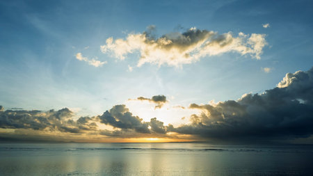 Dramatic sunset over the ocean with golden light piercing through scattered clouds, reflecting on...