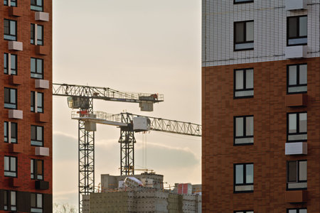 Tower cranes and unfinished building seen between modern apartment blocks at sunset, illustrating...