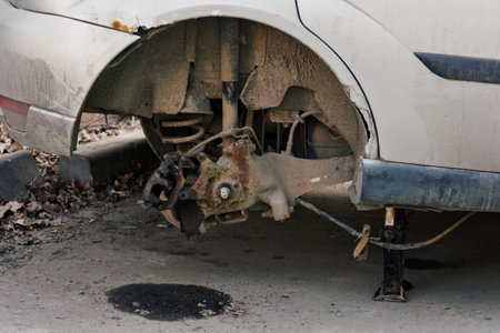 Old car with missing rear wheel stands on jack in urban yard, exposing rusty suspension and dirty undercarriage in neglected condition.の写真素材