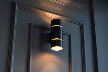 Modern black cylinder wall sconce illuminates a dark paneled wall, creating a warm upward and downward glow with soft shadows and elegant lines.の写真素材