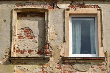 Weathered wall of an old building with peeling plaster, exposed red bricks, one bricked-up window and one modern window with curtain.の写真素材