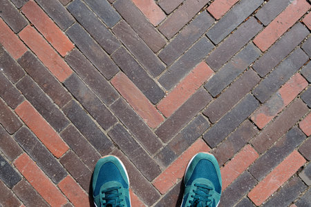 Pair of blue sneakers stands on a herringbone brick pavement, highlighting urban geometry and personal perspective in outdoor environment.の写真素材