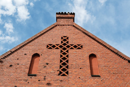 Red brick church facade with geometric cross-shaped window and arched openings, illuminated by soft daylight against blue sky with clouds.の写真素材