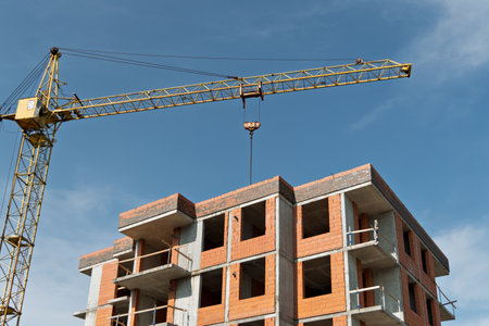 Tower crane rises above unfinished brick apartment building, symbolizing progress and growth in urban construction under a bright blue sky.の写真素材