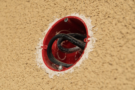 Exposed black electrical wires coiled inside a red plastic mounting box set in textured beige wall during installation or renovation work.の写真素材