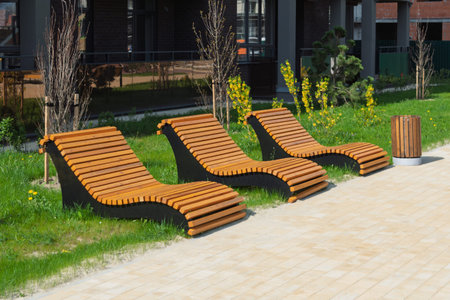 Three modern wooden lounge chairs with curved design stand on green grass in a sunny urban...