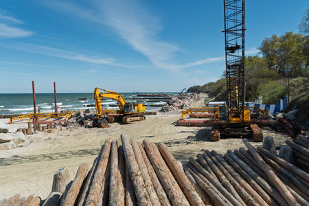 Yellow construction equipment and timber piles at a coastal site, with machinery working on shoreline reinforcement under a clear blue sky.の写真素材