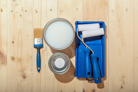 Opened can of clear water-based varnish, brush and rollers with blue tray on wooden floor, showing the start of protective wood finishing.の写真素材