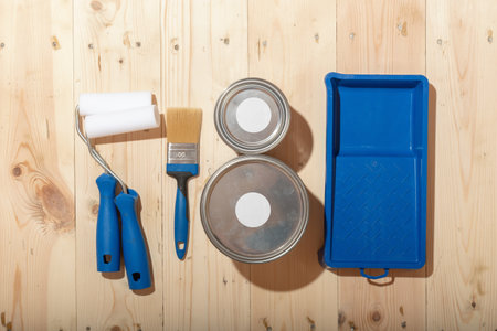 Set of paint rollers, brush, paint cans and blue tray neatly arranged on clean wooden floor, suggesting careful preparation for renovation.の写真素材