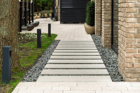 Modern walkway made of concrete slabs leads along landscaped area with decorative gravel and brick building, featuring outdoor lighting.の写真素材