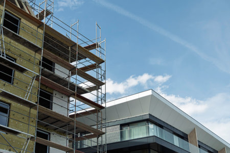 Scaffolding covers building under insulation installation, next to finished modern facade with glass windows and geometric shapes against blue sky.の写真素材