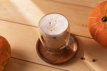Cold pumpkin spice latte with thick foam in double wall glass on wooden table, pumpkins and sunlight enhance warm autumn atmosphereの写真素材