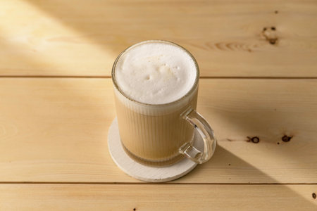 Creamy latte in transparent mug rests on wooden table, soft morning light casting gentle shadows, inviting quiet cozy coffee breakの写真素材