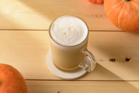 Creamy pumpkin latte in clear glass mug on light wooden table with copy space, cozy autumn drink concept for cafe menu or food blog.の写真素材