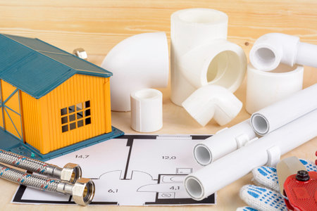 Plumbing project concept with house model, pvc pipes, fittings and hoses on floor plan, symbolizing water system installationの写真素材