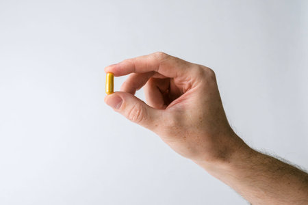 Male hand pinches yellow supplement capsule on white background, calm minimal copy space for...