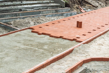 Red paving bricks are laid in grid pattern on prepared base beside concrete steps, showing walkway...