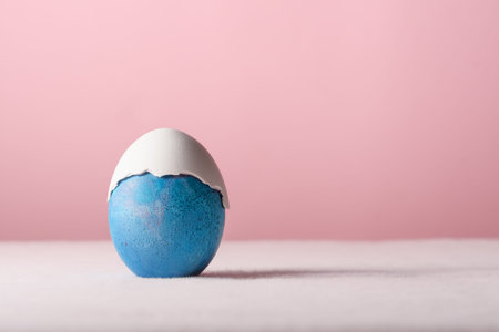 Blue Easter egg wears cracked white shell cap, minimalist pastel still life with gentle humor and...