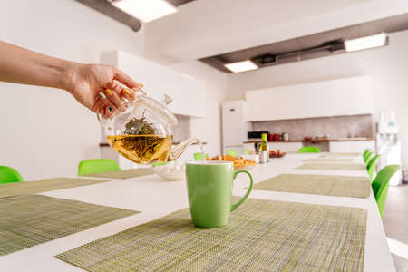 A woman holds in her hand a transparent teapot for tea. Green tea in the teapot. A woman pours green tea into a green cup. Modern cuisine.の写真素材
