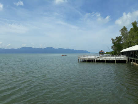 A pontoon on the edge of a mangrove forest with a clear sea and sky. There are a boat and mountains in the back.の写真素材