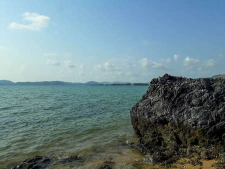 Rough-skinned rocks by the blue sea, the sky is clear, the mountains can be seen in the distance.の写真素材