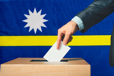A hand holds a ballot paper in a ballot box against the background of the Nauru flag. People place ballot papers in a ballot box with the Nauru flag in the background. Elections in Nauru.の写真素材