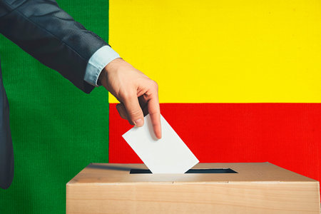 A hand holds a ballot in a ballot box against the background of the Benin flag. People cast ballots...