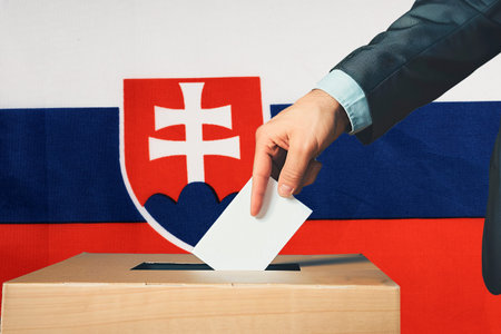 A hand holds a ballot in a ballot box against the background of the Slovakia flag. People cast...