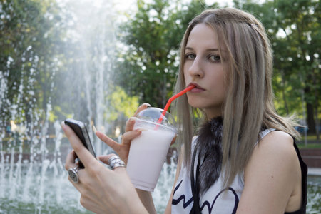 Girl with mobile phone drinks cocktail in the park in front of the fountain