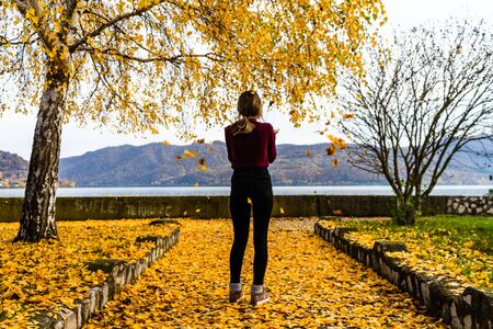 Girl throws up fallen maple leaves. Autumn season, leaves on the ground, river and mountains in the background.の写真素材
