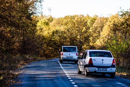 Two cars on autumn road in forest. Autumn season with sunlight. Colorful trees and fallen leaves in autumn forest. Lugoj, Romania, 2019.のeditorial素材