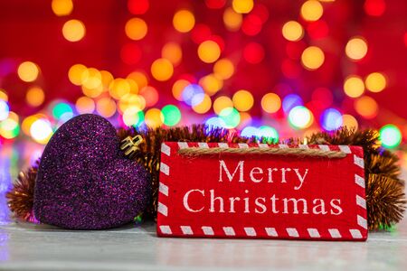 Merry Christmas wooden sign and colorful heart shaped ornament. Christmas composition on blurred lights background.の写真素材