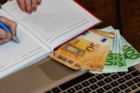 Different objects on wooden office desk. Modern wooden office desk table with laptop keyboard, euro money, office agenda. Business woman writing on office agenda.の写真素材