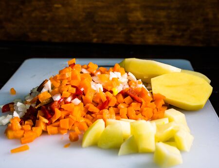 Fresh and frozen chopped vegetables,  Chopped carrots, potatoes, onions and red bell pepper on white cutting board.の写真素材