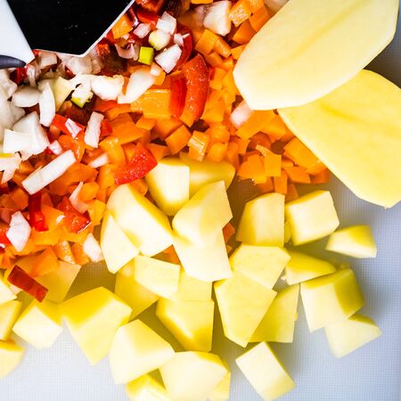 Fresh and frozen chopped vegetables,  Chopped carrots, potatoes, onions and red bell pepper on white cutting board.の写真素材
