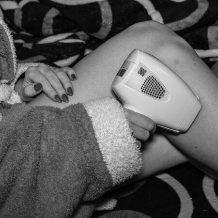 Close up view of woman shaving legs in bed at home. Shaving legs and skin care, girl with a hair laser remover, IPL cosmetology device. Unshaven female legs on the bed, hair removal concept. Bucharest, Romania, 2020.のeditorial素材