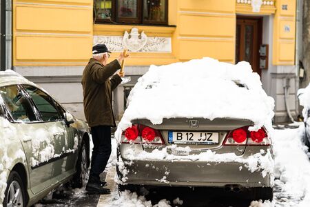 Old man, male in winter coat cleaning, shoveling driveway, street from covered falling snow with shovel, residential houses, cars parked on road in Bucharest, Romania 2020のeditorial素材