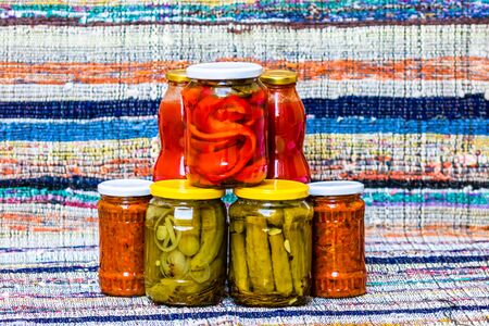 Glass jars with pickled red bell peppers and pickled cucumbers (pickles) isolated. Jars with variety of pickled and canned vegetables. Preserved food concept in a rustic composition.の写真素材