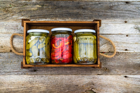 Glass jars with pickled red bell peppers and pickled cucumbers (pickles) isolated in wooden crate....