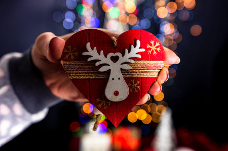 Holding Christmas heart shape decoration isolated on background with blurred lights. December season, Christmas composition.の写真素材