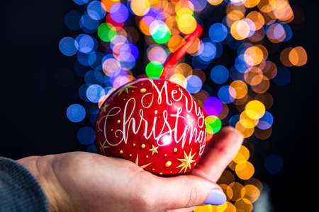 Holding Merry Christmas bauble decoration isolated on background with blurred lights. December season, Christmas composition.の写真素材