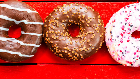 Colorful donuts on red wooden table. Sweet icing sugar food with glazed sprinkles, doughnut with chocolate frosting. Top view with copy spaceの写真素材