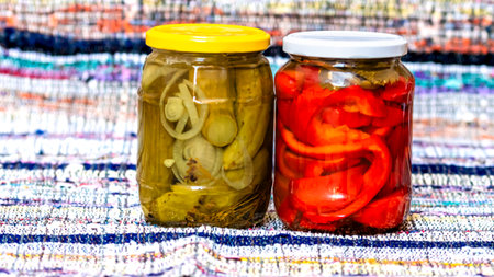 Glass jars with pickled red bell peppers and pickled cucumbers (pickles) isolated. Jars with variety of pickled vegetables. Preserved food concept in a rustic composition.の写真素材
