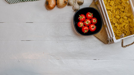 Top view of onions, garlic, pasta in a wooden crate and fresh ripe cherry tomatoes on a rustic white wooden table. Ingredients and food conceptの写真素材